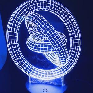 May include: A blue LED lamp with a 3D geometric design. The lamp features a white grid pattern forming a circular shape around a knotted structure. The base has a power symbol. The lamp emits a bright blue glow against a dark background.