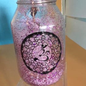 May include: A clear glass jar filled with pink glitter and a black mandala design with an Om symbol. The jar has a purple ribbon around the lid and sits on a clear acrylic stand.