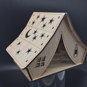 May include: A wooden tent-shaped decorative piece with laser-cut details. The roof features stars and a crescent moon, while the sides have a window and a lantern design. The natural wood color is accented by dark outlines.