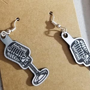 May include: A pair of silver-tone microphone earrings. Each earring features a detailed outline of a vintage microphone design with a stand. The earrings have a hook closure for pierced ears. The background is a light brown color.