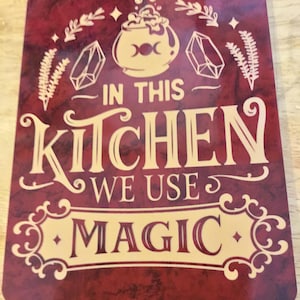 May include: A burgundy kitchen sign with the text "IN THIS KITCHEN WE USE MAGIC" in cream-colored lettering. The sign features a cauldron, crystals, and botanical illustrations.