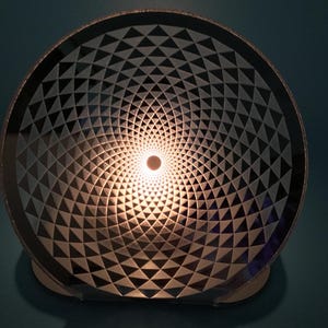 May include: A decorative lamp with a geometric design. The lamp features a circular shade with a pattern of triangles, creating a spiral effect. The center of the lamp emits a warm, inviting light. The base is a light color.