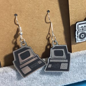 May include: Silver-toned computer earrings with black details. The earrings feature a retro computer design with a screen and keyboard. The earrings hang from silver-colored fish hook ear wires. The earrings are displayed on a brown surface.