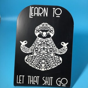 May include: Black sign with white text and a white sloth illustration in a yoga pose. The text reads "LEARN TO" at the top and "LET THAT SHIT GO" at the bottom. The background is a solid blue.