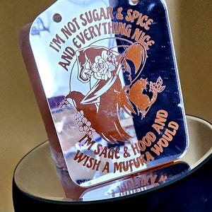 May include: A rectangular, mirrored acrylic tag with a decorative design and text. The design features a witch with floral accents in a copper color. The text reads "I'M NOT SUGAR & SPICE AND EVERYTHING NICE I'M SAGE & HOOD AND WISH A MUFUKA WOULD."