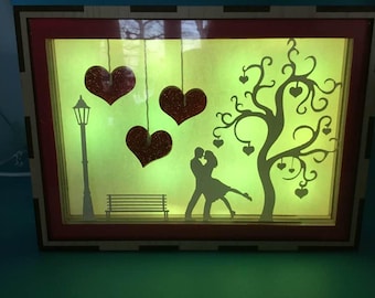 May include: A decorative illuminated box featuring a romantic scene. Silhouettes of a couple, a tree with heart-shaped leaves, a street lamp, and a bench are set against a glowing background. Three glittery red hearts hang from above.