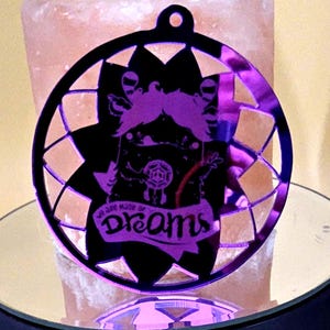 May include: A circular, purple and black ornament with a dreamcatcher design. The ornament features a cartoon figure and the words "We are made of Dreams". The ornament is hanging from a small hole at the top.