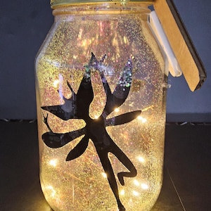May include: A decorative glass jar filled with glitter and fairy lights. A black silhouette of a fairy is inside the jar. The jar has a gold lid and a gold ribbon around the top.