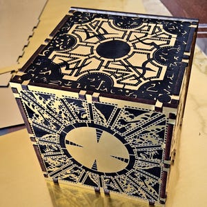 May include: A wooden puzzle box with intricate geometric and symbolic designs. The box features a gold and black color scheme, with detailed patterns etched into the surface. The cube-shaped box is a decorative item.