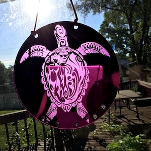 May include: A round, black decorative hanging featuring a pink sea turtle design. The turtle is intricately detailed with tribal patterns. The sun shines through the window, illuminating the pink design. The hanging is suspended by a black cord.