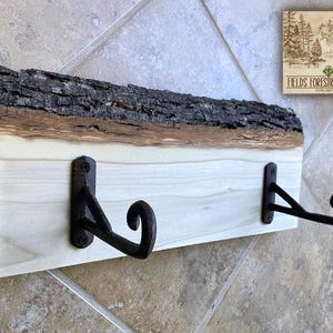 May include: A rustic wooden wall hook with two black metal hooks. The top edge features the natural bark of the tree, adding a unique texture. The wood is light-colored, and the hooks are mounted on the front. The sign in the corner reads "FIELDS FORESTREE FINDS ESTD 2017".