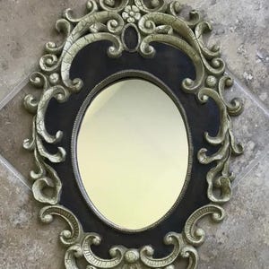 May include: Oval-shaped decorative mirror with an ornate, antique-style frame. The frame is a combination of black and gold, with intricate scrollwork and floral accents. The mirror itself has a light yellow tint.