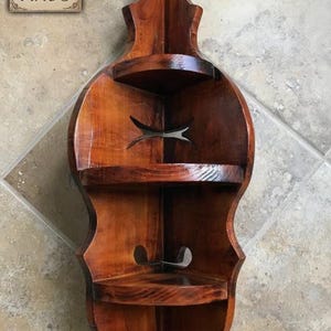 May include: A wooden corner shelf with a violin-like shape, featuring two shelves and decorative cutouts. The shelf is stained a warm brown color and has a glossy finish. The sign in the image reads "FOUR FIELDS FINDS".