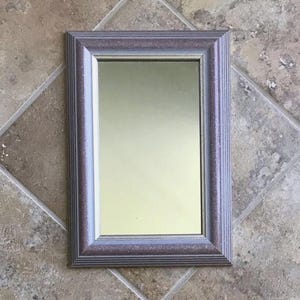 May include: A rectangular wall mirror featuring a textured, dark mauve frame with a subtle metallic sheen. The frame has a multi-layered, stepped profile with fine linear grooves along the outer and inner edges, surrounding a clear glass center.