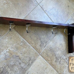 May include: A dark brown wooden wall shelf featuring four silver-colored metal hooks shaped like bent forks. The shelf has two side supports and a flat horizontal surface. A small square logo in the corner displays the text FOUR FIELDS FINDS.