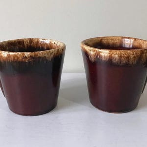 May include: Two vintage brown ceramic mugs with a unique handle design. The mugs have a dark brown base with a lighter brown rim. The handles feature a circular opening. The mugs are displayed on a white surface.