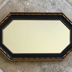 May include: An octagonal mirror with a black frame and gold trim. The mirror's frame has a decorative rope-like design. The mirror's surface is a light cream color, reflecting the surrounding environment.
