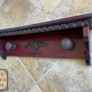 May include: A distressed red wooden shelf with two round knobs and a decorative metal accent. The shelf has a black trim and is mounted on a wall. The shelf is from Four Fields Finds.