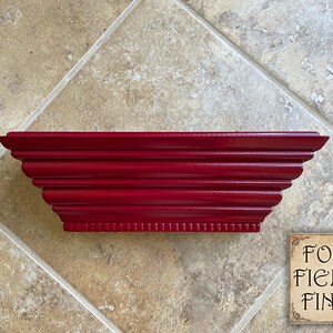 May include: A deep red, rectangular wooden shelf with a decorative, layered design. The shelf has a series of horizontal grooves and a detailed edge. The Four Fields Finds logo is in the lower right corner.