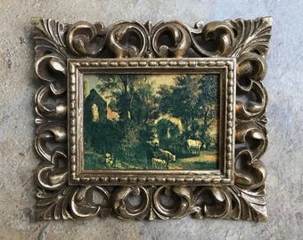 May include: A small framed landscape painting featuring a pastoral scene with cows, trees, and a building. The ornate gold frame has a detailed, scrollwork design. The painting's colors include greens, browns, and yellows.