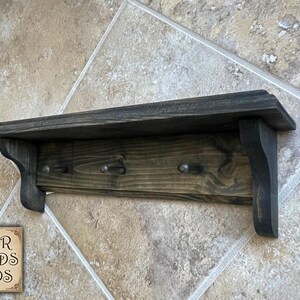 May include: A dark stained wooden shelf with three metal hooks. The shelf has decorative curved supports and is mounted on a tiled wall. The sign in the corner reads "FOUR FIELDS FINDS".