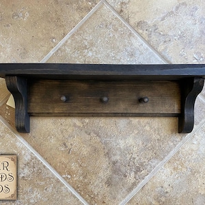 May include: A dark stained wooden shelf with three pegs below. The shelf has a decorative edge and is supported by two curved brackets. The sign in the lower left corner reads "FOUR FIELDS FINDS".
