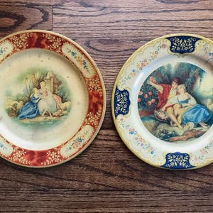May include: Two decorative plates with romantic scenes. One plate has a red and floral border with a scene of figures in a garden. The other plate has a blue and floral border with a couple in a garden setting.