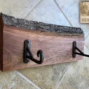 May include: A rustic wooden wall-mounted coat rack with two black metal hooks. The wood has a natural edge with bark, and the hooks are attached with screws. The sign in the corner reads "FIELDS FORESTREE FINDS".