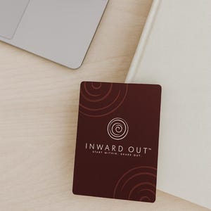 Peut inclure: Une carte de visite marron avec le texte "INWARD OUT" et le slogan "START WITHIN. SHARE OUT." est affich&eacute;e sur une surface en bois clair. Un ordinateur portable et un livre blanc sont &eacute;galement visibles.