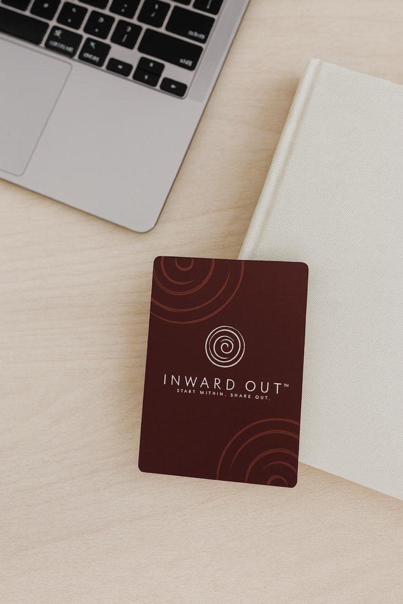 Peut inclure: Une carte de visite marron avec le texte "INWARD OUT" et le slogan "START WITHIN. SHARE OUT." est affich&eacute;e sur une surface en bois clair. Un ordinateur portable et un livre blanc sont &eacute;galement visibles.