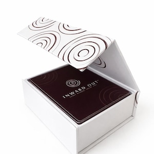 Peut inclure: Une bo&icirc;te blanche avec un couvercle &agrave; charni&egrave;re contenant un jeu de cartes. Le couvercle et le dessus des cartes pr&eacute;sentent un motif en spirale marron. Les cartes sont marron fonc&eacute; avec le texte "INWARD OUT" et un logo en spirale.
