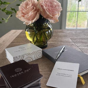 Peut inclure: Nature morte avec un vase contenant des roses roses, une bo&icirc;te et des cartes "INWARD OUT", un journal gris et un stylo. Une carte affiche "VALUES & BOUNDARIES". L'ensemble est pos&eacute; sur une table en bois.