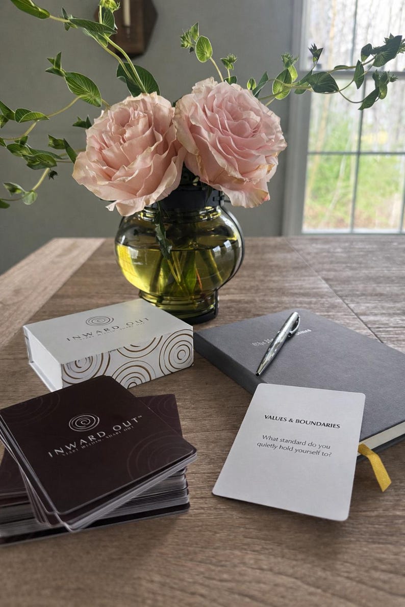 Peut inclure: Nature morte avec un vase contenant des roses roses, une bo&icirc;te et des cartes "INWARD OUT", un journal gris et un stylo. Une carte affiche "VALUES & BOUNDARIES". L'ensemble est pos&eacute; sur une table en bois.