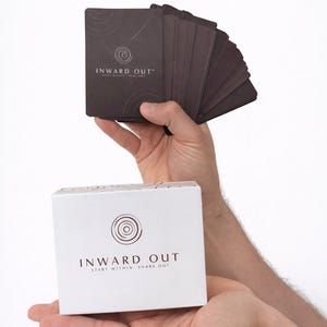 Peut inclure: Un jeu de cartes brun fonc&eacute; et une bo&icirc;te blanche sont tenus en mains. Les cartes et la bo&icirc;te pr&eacute;sentent le texte "INWARD OUT" avec un motif circulaire. La bo&icirc;te comprend &eacute;galement le texte "START WITHIN SHARE OUT". Les cartes semblent &ecirc;tre pour un jeu ou une m&eacute;ditation.