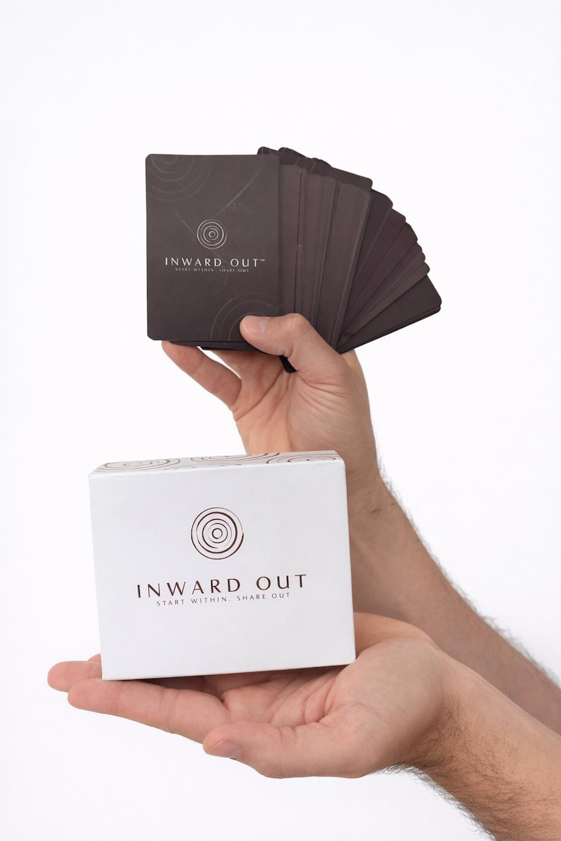 Peut inclure: Un jeu de cartes brun fonc&eacute; et une bo&icirc;te blanche sont tenus en mains. Les cartes et la bo&icirc;te pr&eacute;sentent le texte "INWARD OUT" avec un motif circulaire. La bo&icirc;te comprend &eacute;galement le texte "START WITHIN SHARE OUT". Les cartes semblent &ecirc;tre pour un jeu ou une m&eacute;ditation.