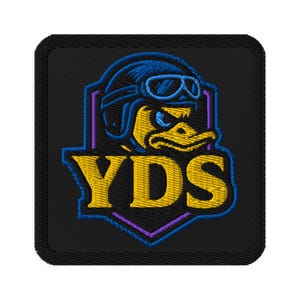 May include: Embroidered patch featuring a duck wearing a blue aviator helmet and goggles. The duck is yellow with blue accents, set against a black background. The letters "YDS" are in yellow. The patch has a purple and blue border.