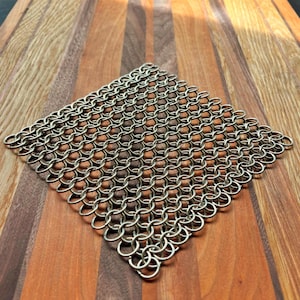 Può includere: Una spugna a catena di metallo argentata, composta da anelli metallici interconnessi. La spugna è posizionata diagonalmente su un tagliere di legno con un motivo a strisce. Gli anelli metallici sono fittamente intrecciati.