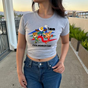 May include: A heather gray short-sleeve t-shirt featuring a graphic print of three cartoon animal mascots, a soccer ball, the text 26, and the words FIFA WORLD CUP. The shirt is paired with dark blue denim jeans with a visible metal button and rivets.