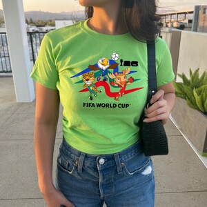 May include: A lime green short-sleeve t-shirt featuring a graphic print of three cartoon animal mascots, a soccer ball, and the text 126 FIFA WORLD CUP. The shirt is paired with blue denim jeans and a black textured shoulder bag.
