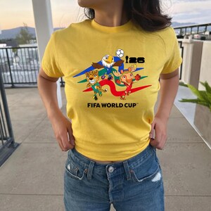 May include: A bright yellow short-sleeve t-shirt featuring a graphic print of three cartoon characters, a soccer ball, and the text 26 FIFA WORLD CUP. The design includes colorful swooshes in blue, green, and red behind the characters.