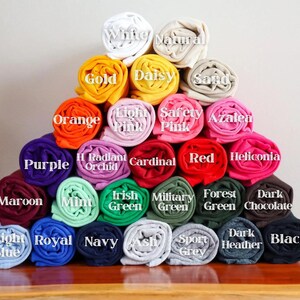 May include: A pyramid stack of rolled fabric swatches in various colors, each labeled with text including White, Natural, Gold, Daisy, Sand, Orange, Light Pink, Safety Pink, Azalea, Purple, Radiant Orchid, Cardinal, Red, Heliconia, Maroon, Mint, Irish Green, Military Green, Forest Green, Dark Chocolate, Light Blue, Royal, Navy, Ash, Sport Grey, Dark Heather, and Black.