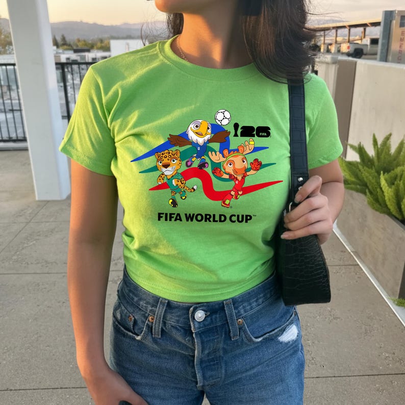 May include: A lime green short-sleeve t-shirt featuring a graphic print of three cartoon animal mascots, a soccer ball, and the text 26 FIFA WORLD CUP. The shirt is paired with blue denim jeans and a black textured shoulder bag with a silver buckle.