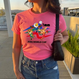 May include: A bright pink short-sleeved t-shirt featuring a graphic print of three cartoon animals, a soccer ball, and the text 26 FIFA WORLD CUP. The design includes blue, green, and red swooshes behind the characters on the front of the shirt.