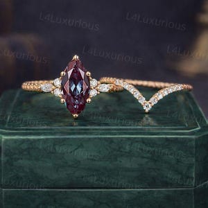 Peut inclure: Une bague de fiançailles en or avec une alexandrite de coupe marquise et une alliance assortie sertie de diamants. L'alexandrite change de couleur, apparaissant bordeaux et sarcelle. La bague repose sur un écrin de velours vert.