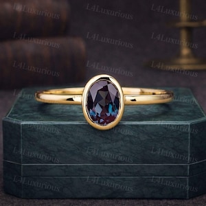 Könnte beinhalten: Ein goldener Ring mit einem oval geschliffenen, dunkelvioletten Edelstein. Der Ring liegt auf einer dunkelgrünen sechseckigen Schatulle. Das Band ist schmal und poliert. Der Hintergrund umfasst verschwommene Bücher und ein dekoratives Objekt.