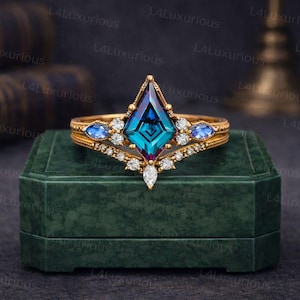 Könnte beinhalten: Ein goldfarbener Ring mit einem großen, teal-farbenen Edelstein in Drachenform, umgeben von kleineren blauen und weißen Steinen. Der Ring wird auf einer dunkelgrünen Samtbox präsentiert.