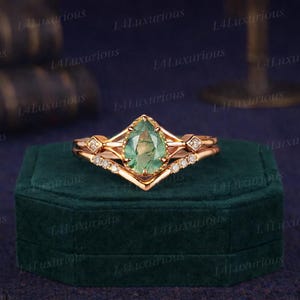 Peut inclure: Un ensemble de bagues en or rose avec une pierre verte en forme de poire et de petits pierres blanches. La bague est sur un écrin de velours vert foncé, avec un fond bleu foncé. La bague a un design unique.