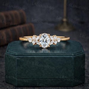 Könnte beinhalten: Ein goldener Ring mit einem oval geschliffenen Mittelstein und kleineren, blütenblattförmigen Steinen auf beiden Seiten. Der Ring liegt auf einer dunkelgrünen Schmuckschatulle, die sein elegantes Design hervorhebt.