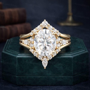 Könnte beinhalten: Ein dreiteiliges goldenes Ringset. Der Verlobungsring verfügt über einen großen, oval geschliffenen Diamanten, der von einem Diamanten-Halo umgeben ist. Der Ring wird von zwei zusätzlichen Goldbändern flankiert, jedes mit kleineren Diamanten. Die Ringe werden auf einer dunkelgrünen Schachtel präsentiert.