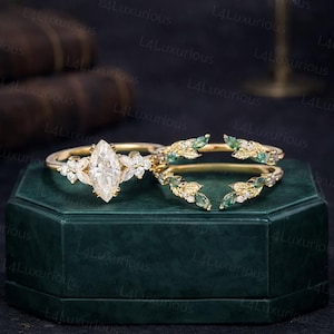 Könnte beinhalten: Ein goldfarbenes Verlobungsring-Set mit einem Mittelstein im Marquise-Schliff und floralen Details. Das Set beinhaltet ein passendes Band mit grünen und weißen Edelsteinen. Die Ringe werden auf einer dunkelgrünen Oberfläche präsentiert.